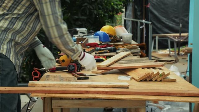 Carpenter wearing gloves arranging stacked wooden planks of different sizes on workbench in workshop
