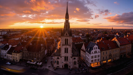 Aerial view of a church with a tall spire at sunset over a European town with red rooftops and orange sky