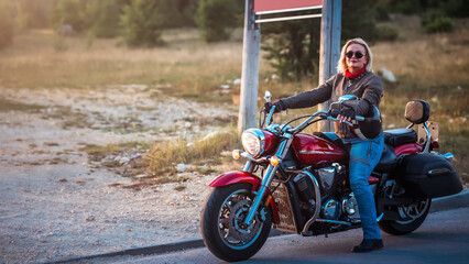 Mature female biker sitting on a red cruiser motorcycle in nature at golden hour, confident and...
