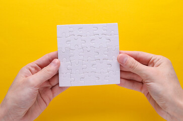 Hand holding white blank assembled square jigsaw puzzle on yellow background. Mockup for design