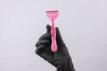 Hand of a cosmetologist in a black glove holds a razor on a gray background. Depilation, epilation,...