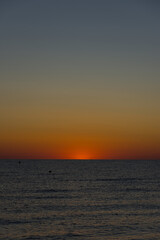 The sun dips below the horizon, casting an orange glow across the tranquil ocean with small buoys...