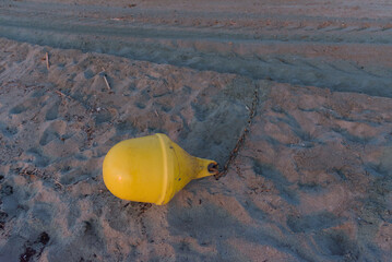 A bright yellow buoy with a chain attached lies on a sandy beach, showing tire tracks in the...
