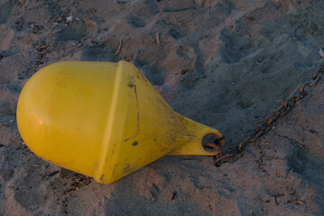 A bright yellow buoy with a chain attached lies on a sandy beach, showing tire tracks in the background.