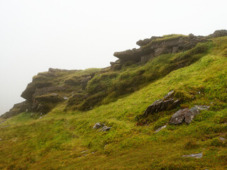 Green grass covers a rocky mountainside as fog rolls in, obscuring the view at Coomloughra...