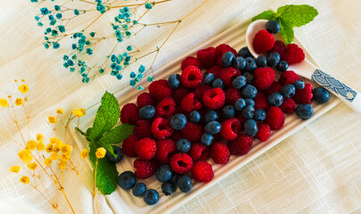 Fresh blueberries and raspberries arranged on a white ceramic tray with mint and colorful dried flowers. Bright and vibrant flat lay for food styling or healthy living themes.

