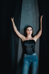A young woman wearing a black corset and blue jeans poses confidently with arms raised.
