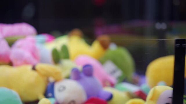 A variety of brightly colored plush toys are displayed inside a claw machine. The toys sit behind a clear glass barrier in an indoor arcade environment, ready to be won by players.