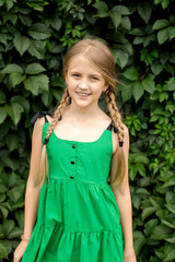 A blonde girl with pigtails in a green sundress is smiling against a leafy background.