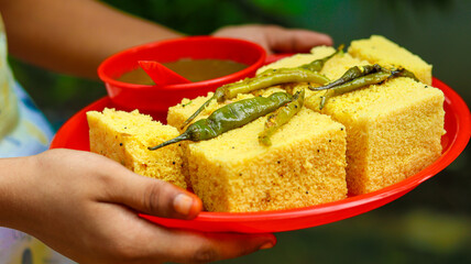 Khaman dhokla served on a red plate with green chutney in a red bowl, garnished with green chillies. Held in hand ,A vibrant and tasty Indian snack presentation, selective focus 