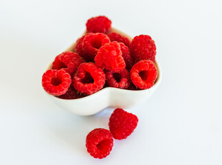 Close-up of fresh red raspberries in a white ceramic heart-shaped bowl on white background. Minimal, clean and romantic food concept.