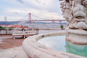 A stone fountain with a sculpted fish head in the foreground, overlooking a cityscape with a large suspension bridge in the background. The sky is cloudy, and a person sits on a bench nearby.