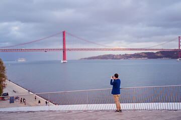 A person taking a photo of a red suspension bridge over a river on a cloudy day.
