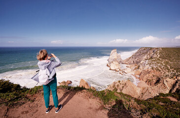 A person with a backpack takes a photo of a scenic coastal view with cliffs and ocean waves.