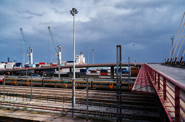 Fototapeta premium A busy industrial port with cranes, shipping containers, and railway tracks under a cloudy sky.