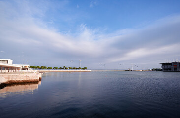 Calm body of water with a cable car system overhead. The sky is partly cloudy, and there are trees lining the waterfront. A pier extends into the water, creating a tranquil atmosphere.