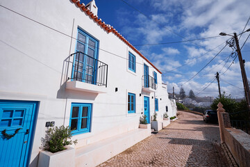 A charming white house with blue accents, featuring balconies and a cobblestone path. The scene is...