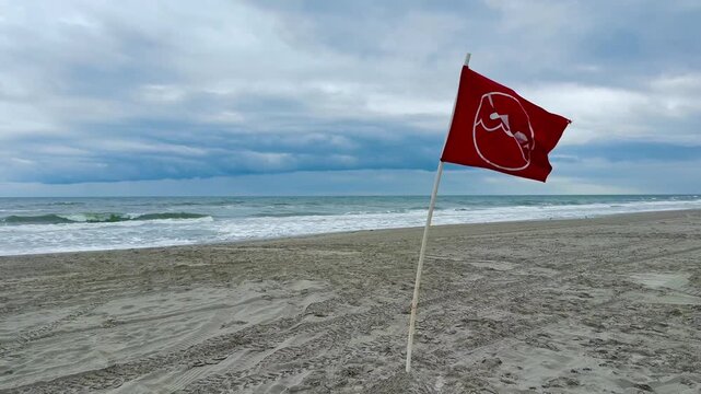 Red flag no swimming warning on a pole in the beach sand near a rough ocean with dark storm clouds on the horizon.