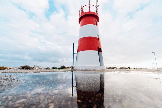 A red and white striped lighthouse stands tall against a cloudy sky, reflected in a puddle on the ground.