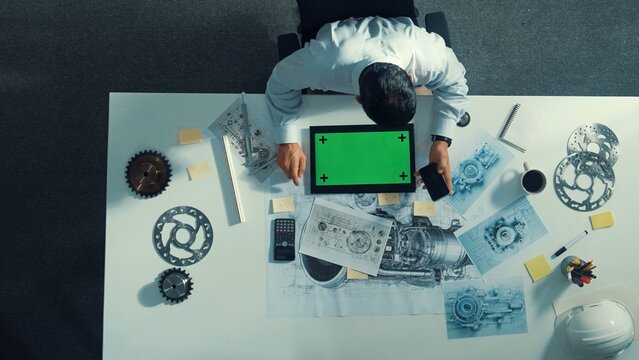Top view of engineer looking at phone while working on tablet with green screen at meeting table with turbine engine or jet engine sketch design and various metal gear scatter around. Alimentation. - Powered by Adobe