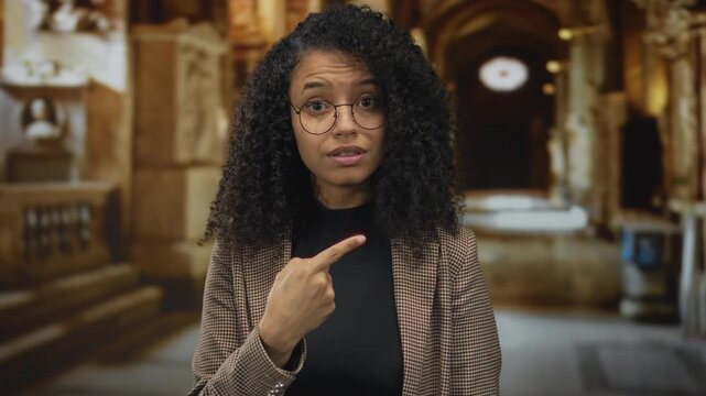 Young hispanic woman wearing round glasses and plaid blazer pointing index finger to herself in ornate church interior; denial.