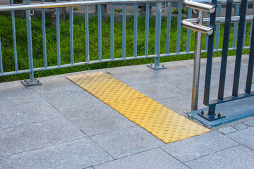 Yellow tactile paving strips provide accessibility guidance along urban walkway with metal safety...