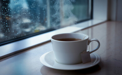 Peacful relaxing at home concept. A hot tea cup with hot steam over cup near by the window glass with water drop,  blurry cityscape in a rainy day through the window background.