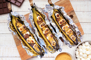 Summer campfire banana smores. Top down view table scene with a white wood background.