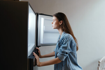 Young woman looking into an open fridge, pondering healthy food choices, with a thoughtful...