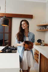 Young woman in a denim shirt and white dress smiling while holding a wooden bowl in a bright...