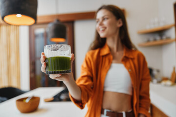 Cheerful young woman in an orange shirt holding a green smoothie in a bright kitchen, symbolizing...