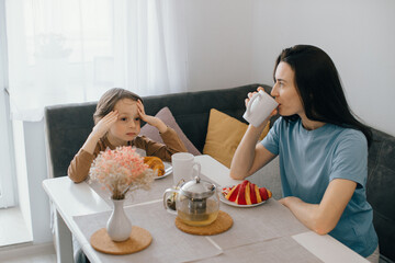 Sad child having breakfast with mother drinking tea at home