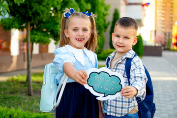 back to school. A girl and a boy students at the school hold a sign with the inscription For the first time in the first grade, a brother and sister with briefcases rejoice at the first school day