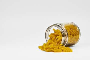 Close up view of raw fusilli pasta (screw pasta) spilling out of a glass container on a white background with copy space.