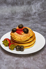 A plate of delicious pancakes in a tower with powdered sugar, decorated with blueberries and fresh berries (strawberries) for a wonderful breakfast.