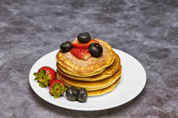 A plate of delicious pancakes in a tower with powdered sugar, decorated with blueberries and fresh berries (strawberries) for a wonderful breakfast.
