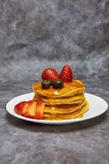 A plate of delicious pancakes in a tower with powdered sugar, decorated with blueberries and fresh berries (strawberries) drizzled with delicious maple syrup for a wonderful breakfast.