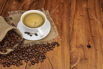 A jute bag with roasted coffee beans scattered on a rustic wooden table, next to it is a cup of freshly brewed coffee