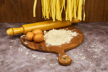 Fresh egg fettuccine noodles, drying in the foreground, hanging over a wooden table containing flour and egg, basic ingredients for preparation.