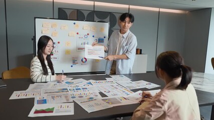 Young asian businessman presenting a financial report to colleagues during a team meeting in a modern office, surrounded by data charts and graphs for project planning and collaboration - Powered by Adobe