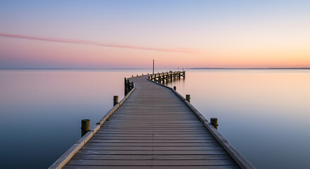Obraz premium A tranquil seascape featuring a long wooden pier stretching towards the horizon over calm, reflective water during a beautiful pastel-colored sunset.