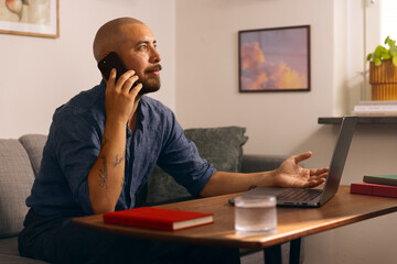 Man working from home on a laptop while holding a smartphone, seated in a cozy living room with books and a glass of water on the table.