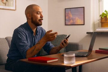 Man working from home on a laptop while holding a smartphone, seated in a cozy living room with books and a glass of water on the table.