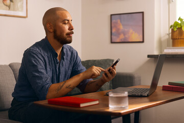Man working from home on a laptop while holding a smartphone, seated in a cozy living room with books and a glass of water on the table.