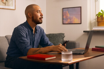 Man working from home on a laptop while holding a smartphone, seated in a cozy living room with...