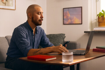 Man working from home on a laptop while holding a smartphone, seated in a cozy living room with...