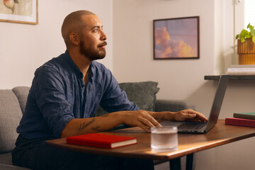 Man working from home on a laptop while seated in a cozy living room with books and a glass of...