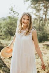 Smiling blonde woman holding a straw hat, enjoying nature in a forest