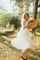 Happy woman wearing white dress and holding straw hat, enjoying sunny day in nature