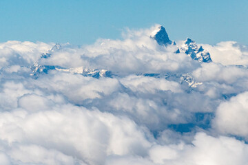 Mid-Air view of Grand Teton's jagged summit emerges above a blanket of fluffy, white clouds under a clear blue sky. The prominence of the peak amidst the clouds outside my airplane window.